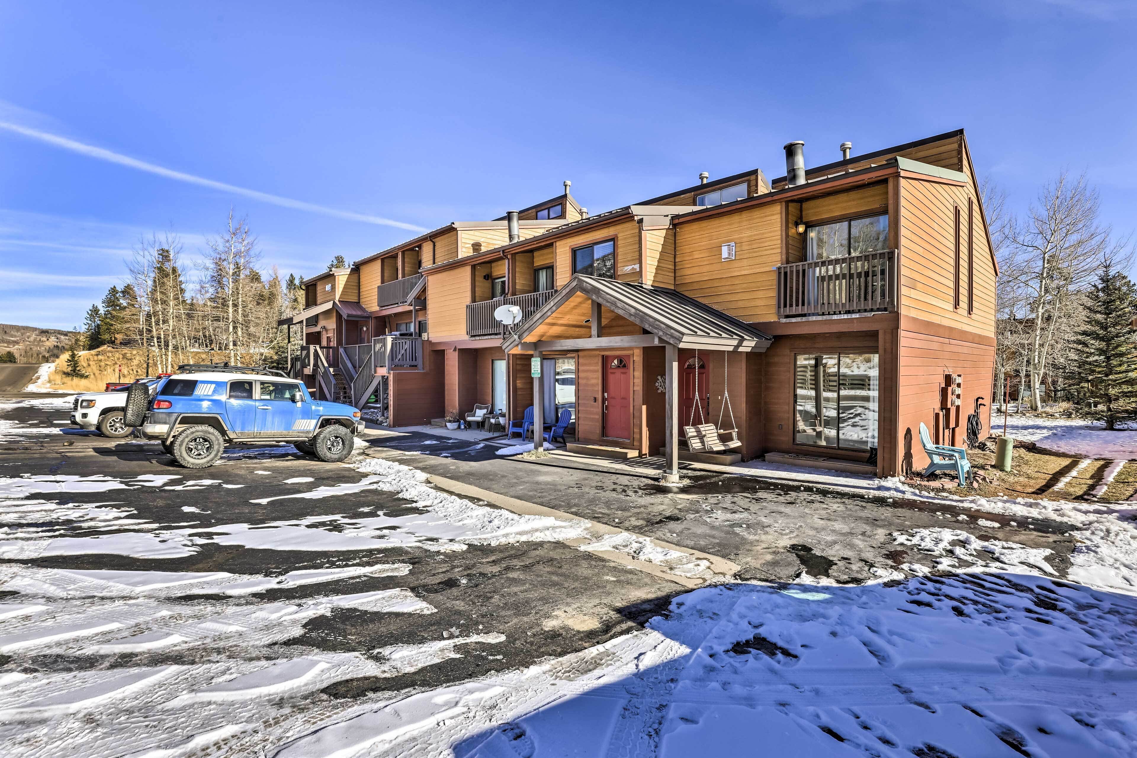 Un immeuble à plusieurs étages doté d’un espace extérieur couvert, un VUS bleu garé devant, et de la neige au sol.