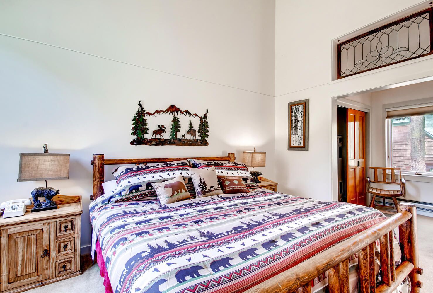 A bedroom with a bed featuring a patterned bedspread, wooden headboard, and a nightstand with a lamp and a phone.