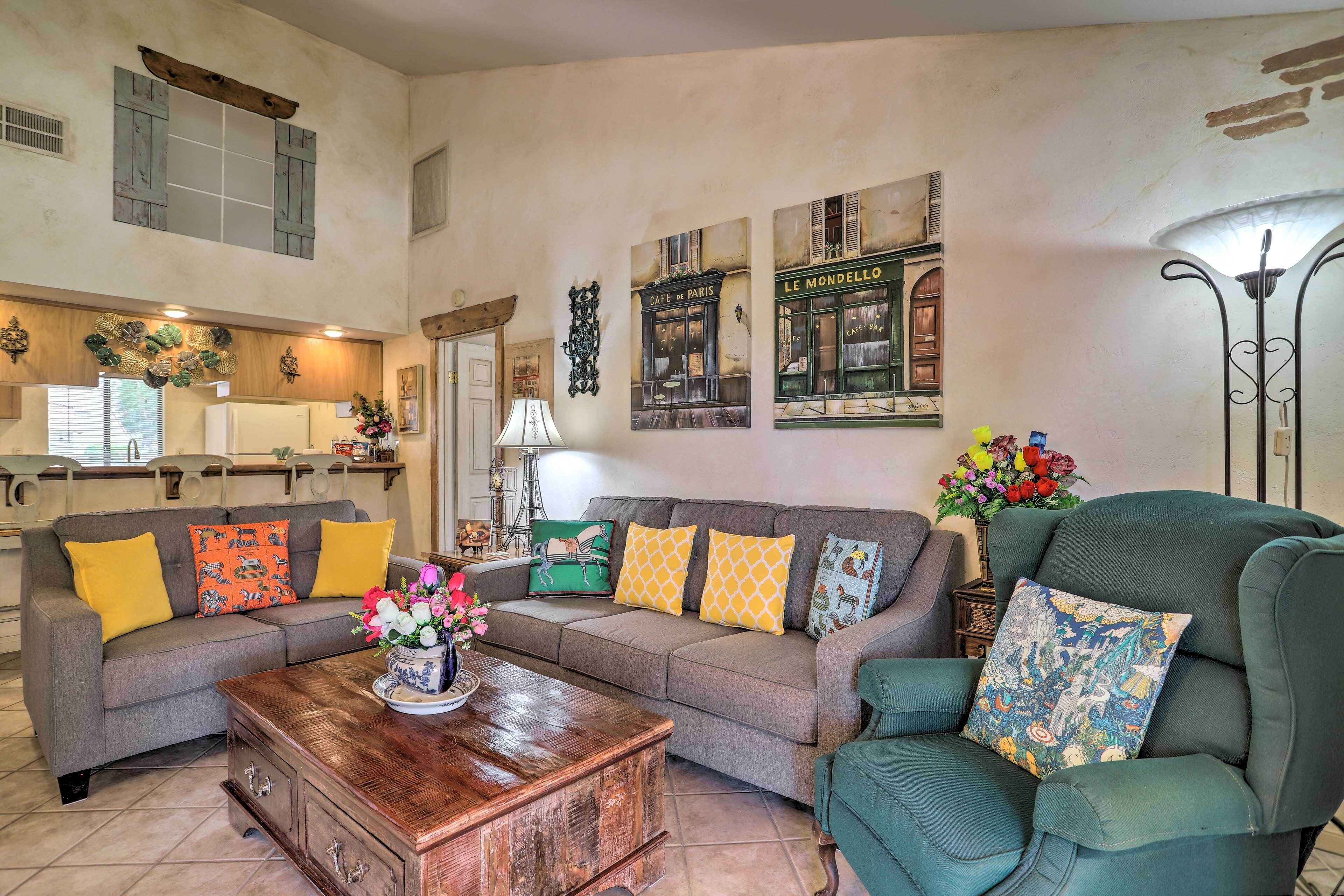 A cozy living room with a grey sofa, a wooden coffee table, and a green armchair. There are decorative pillows and wall art depicting Parisian cafes.