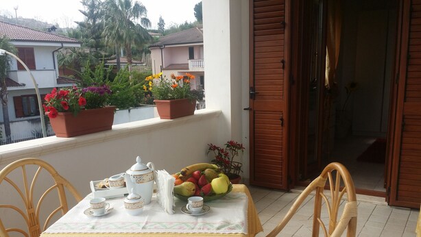 A balcony with a table set for breakfast, potted plants, and a view of neighboring houses.