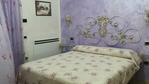 A bedroom with a bed, floral-patterned bedding, a decorative headboard, a wall-mounted picture, and a curtain.