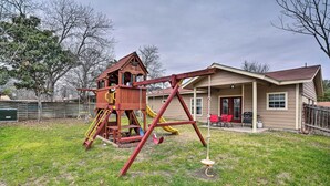 Une structure de jeu en bois avec un toboggan et des balançoires, une maison avec un porche et une cour clôturée.
