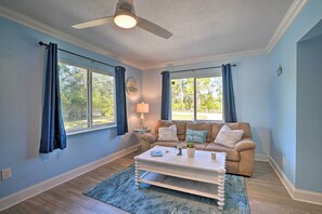 A living room with a beige sofa, a white coffee table, and a blue rug. There are two windows with blue curtains, a ceiling fan, and a lamp on a side table.
