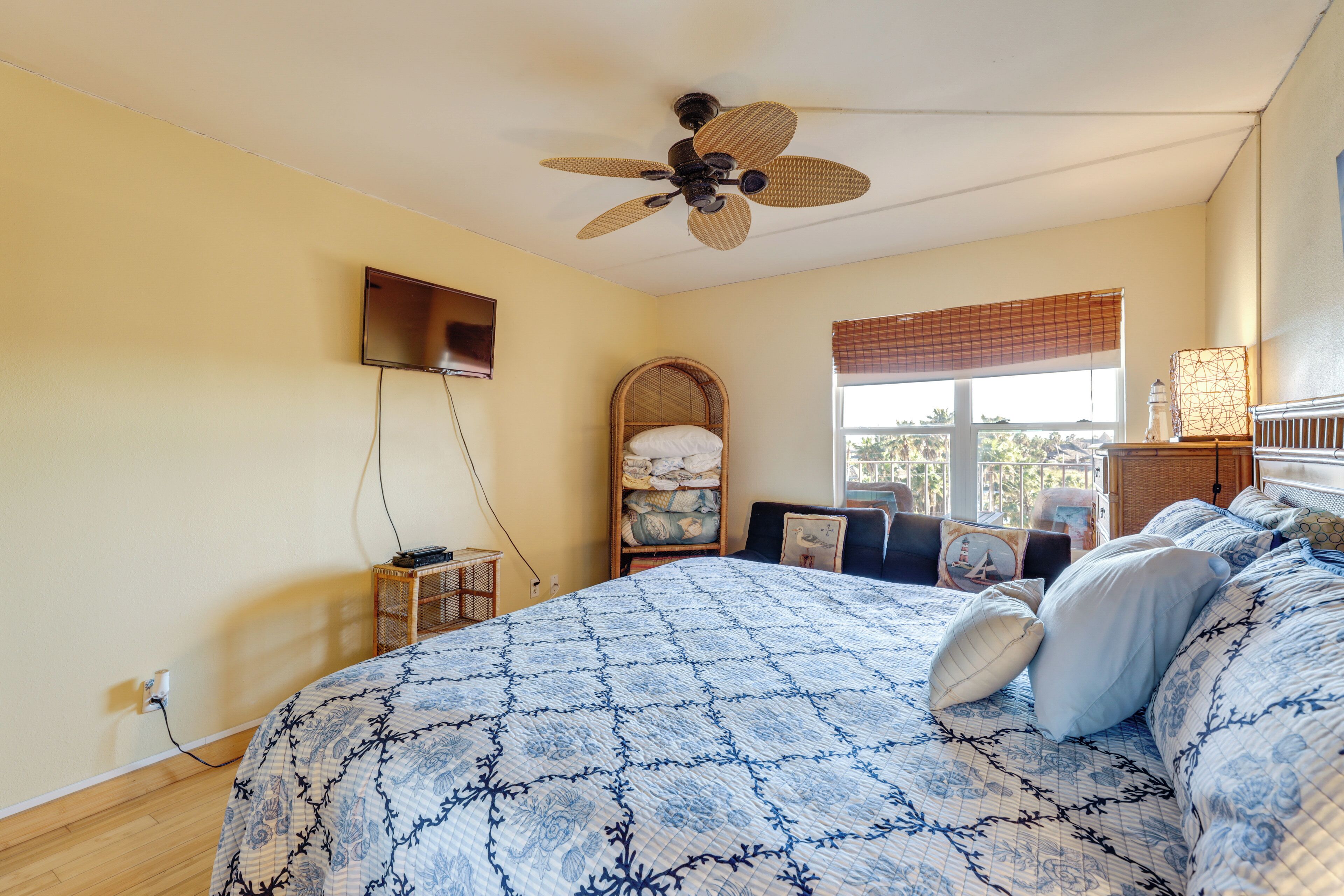 A bedroom with a bed, a ceiling fan, a window with blinds, a bedside table, and a wall-mounted TV.