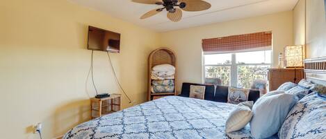 A bedroom with a bed, a ceiling fan, a window with blinds, a bedside table, and a wall-mounted TV.
