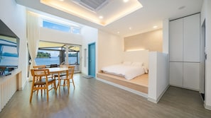 Quarto moderno com cama, área de refeições com cadeiras, vista para a água e uma janela grande.