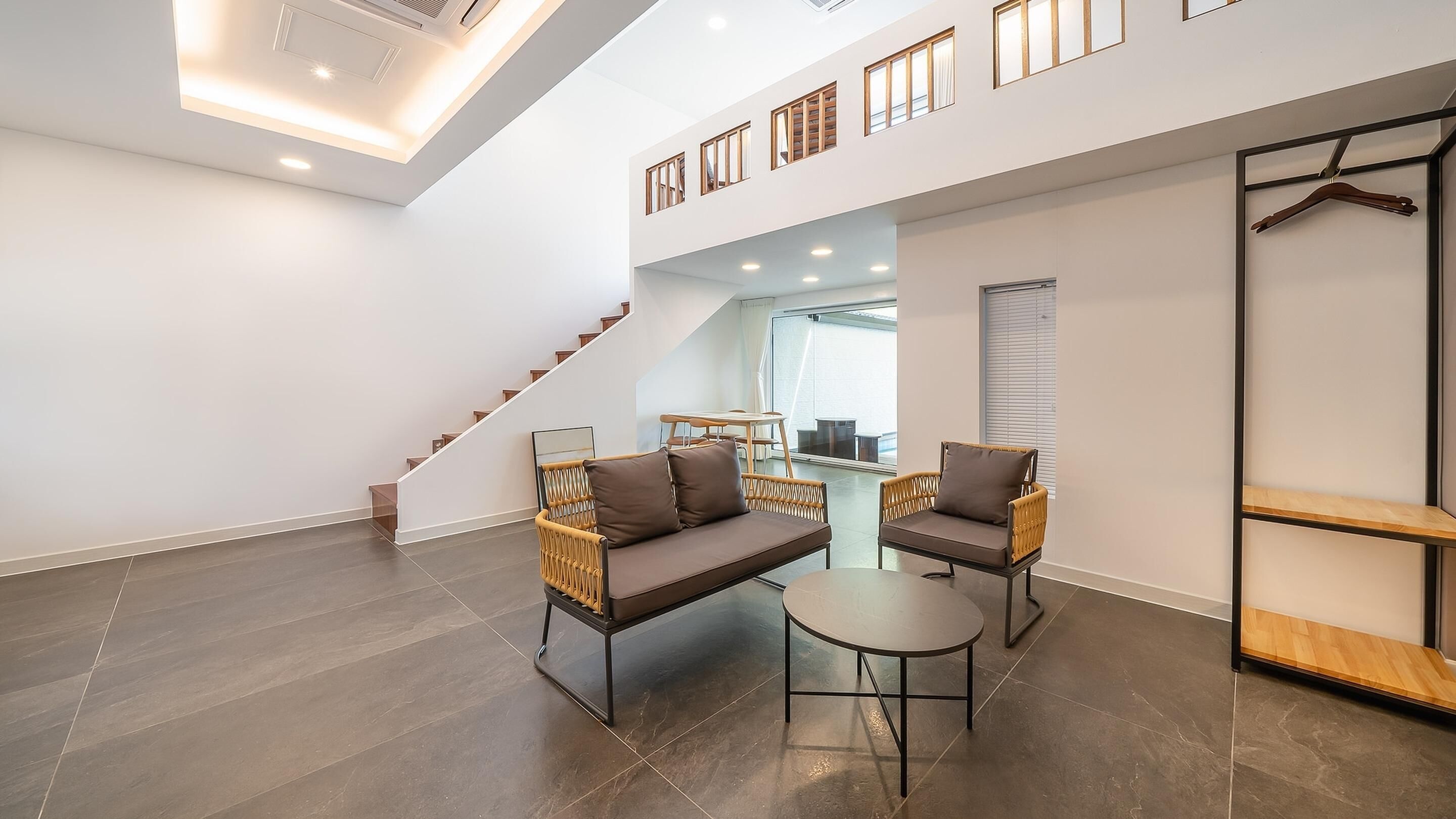 A spacious living area with a high ceiling, a staircase, a sofa set, a round table, and a wooden bench.