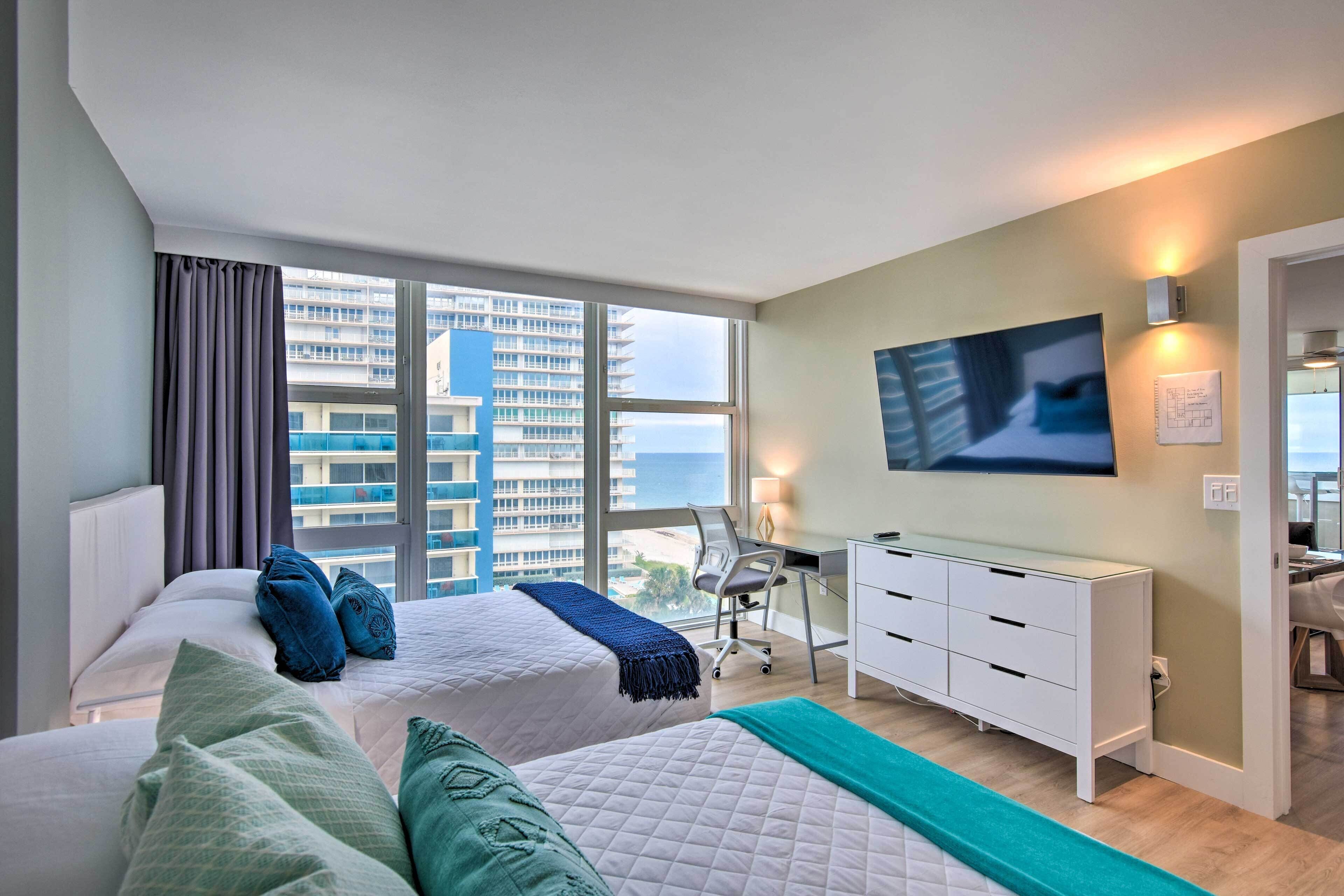Ein Hotelzimmer mit zwei Betten, einem Schreibtisch, einem Sessel, einem Fernseher und Blick auf die Stadt.