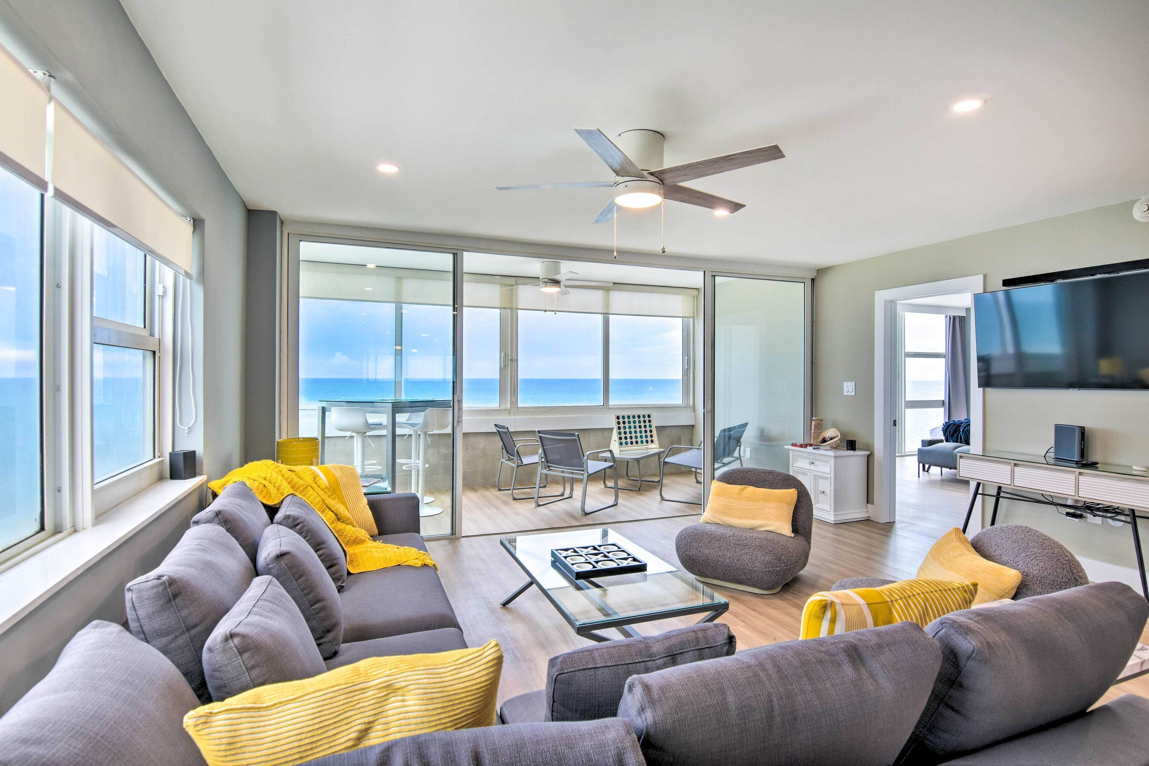 A modern living room with a sofa, coffee table, and a TV. There is a balcony with a view of the ocean.