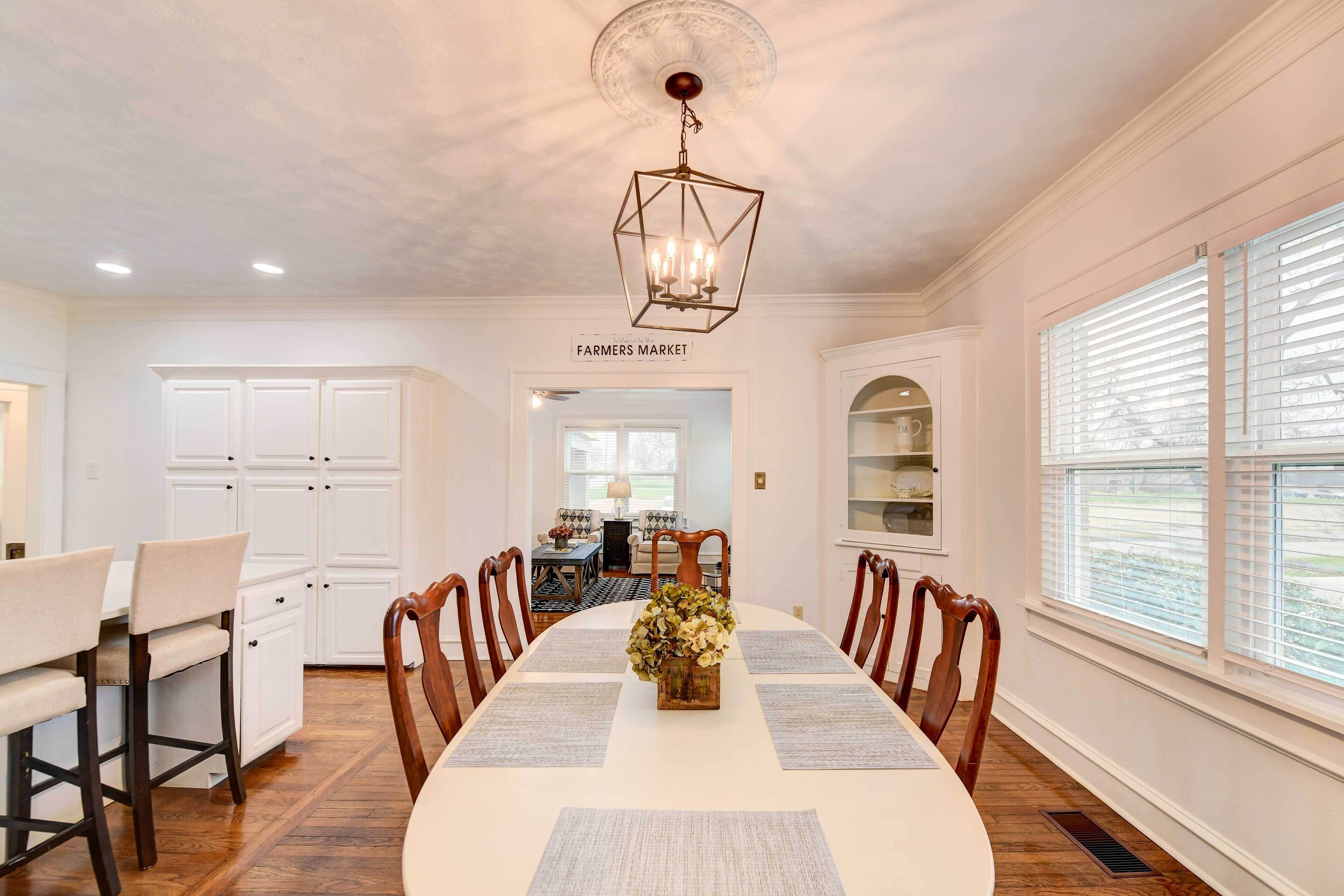 A dining room with a long table, chairs, and a chandelier. There is a kitchen area with white cabinets and a window with blinds.