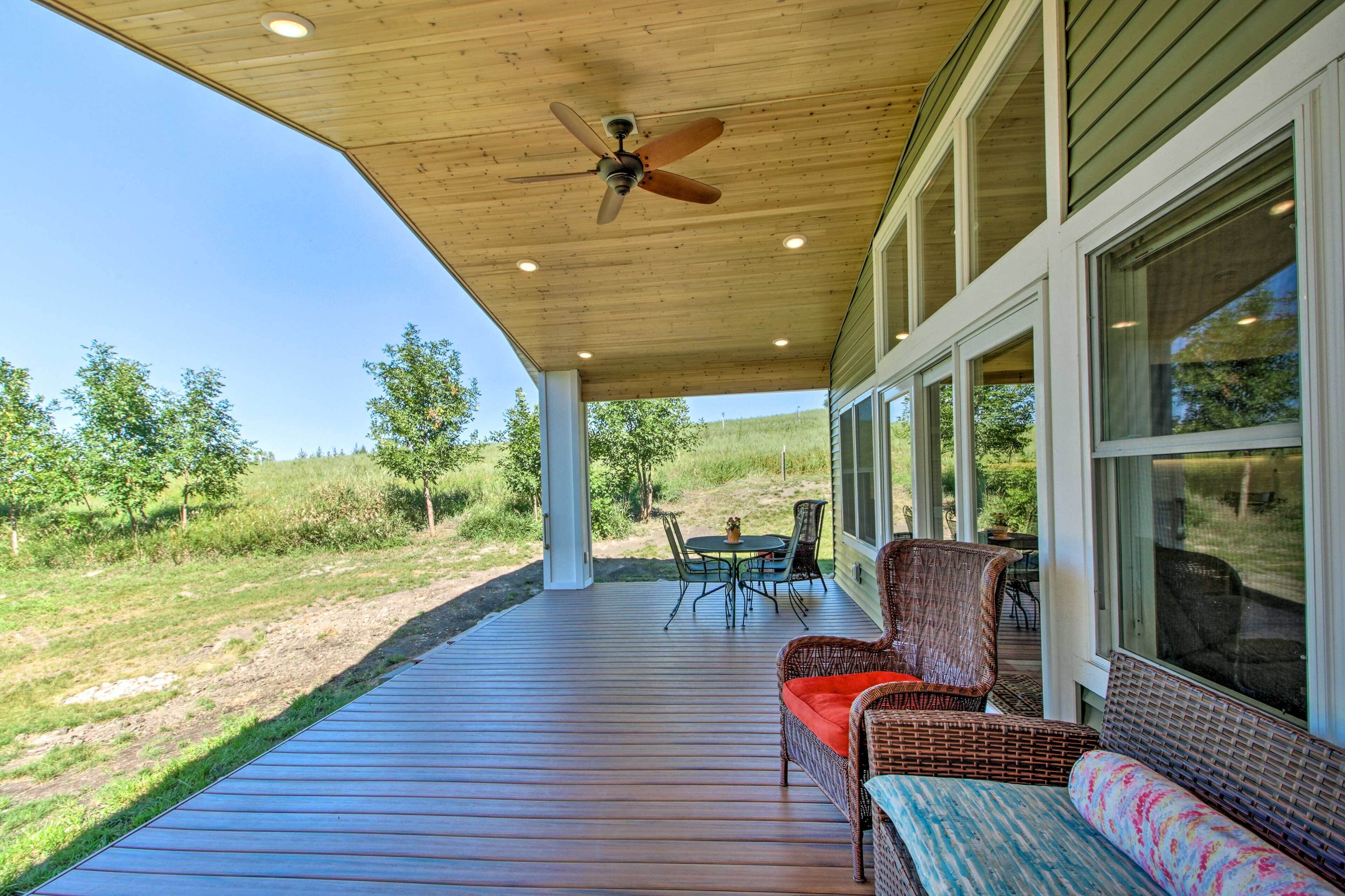 A covered outdoor deck with a ceiling fan, wicker furniture, and large windows overlooking a grassy area.