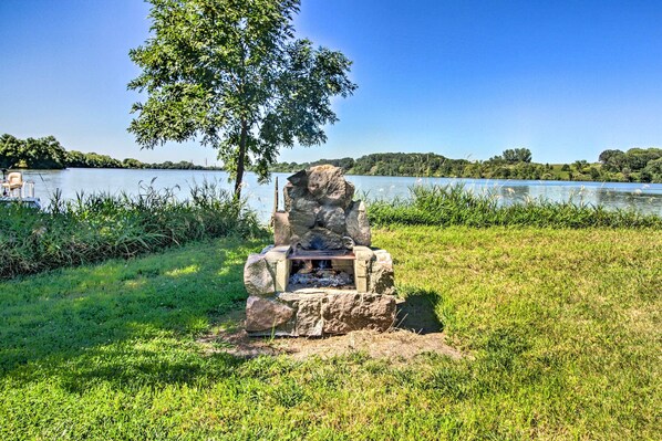 A stone fire pit surrounded by grass and a tree, with a lake and boats in the background.