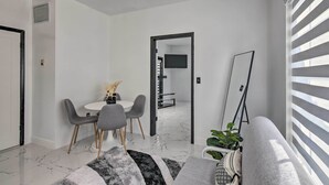 Un salon moderne comprenant un canapé, une table à manger avec des chaises, un téléviseur à écran plat fixé au mur et une grande fenêtre avec des stores.