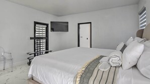 Une chambre avec un grand lit, un téléviseur fixé au mur, une porte et une fenêtre avec des stores.