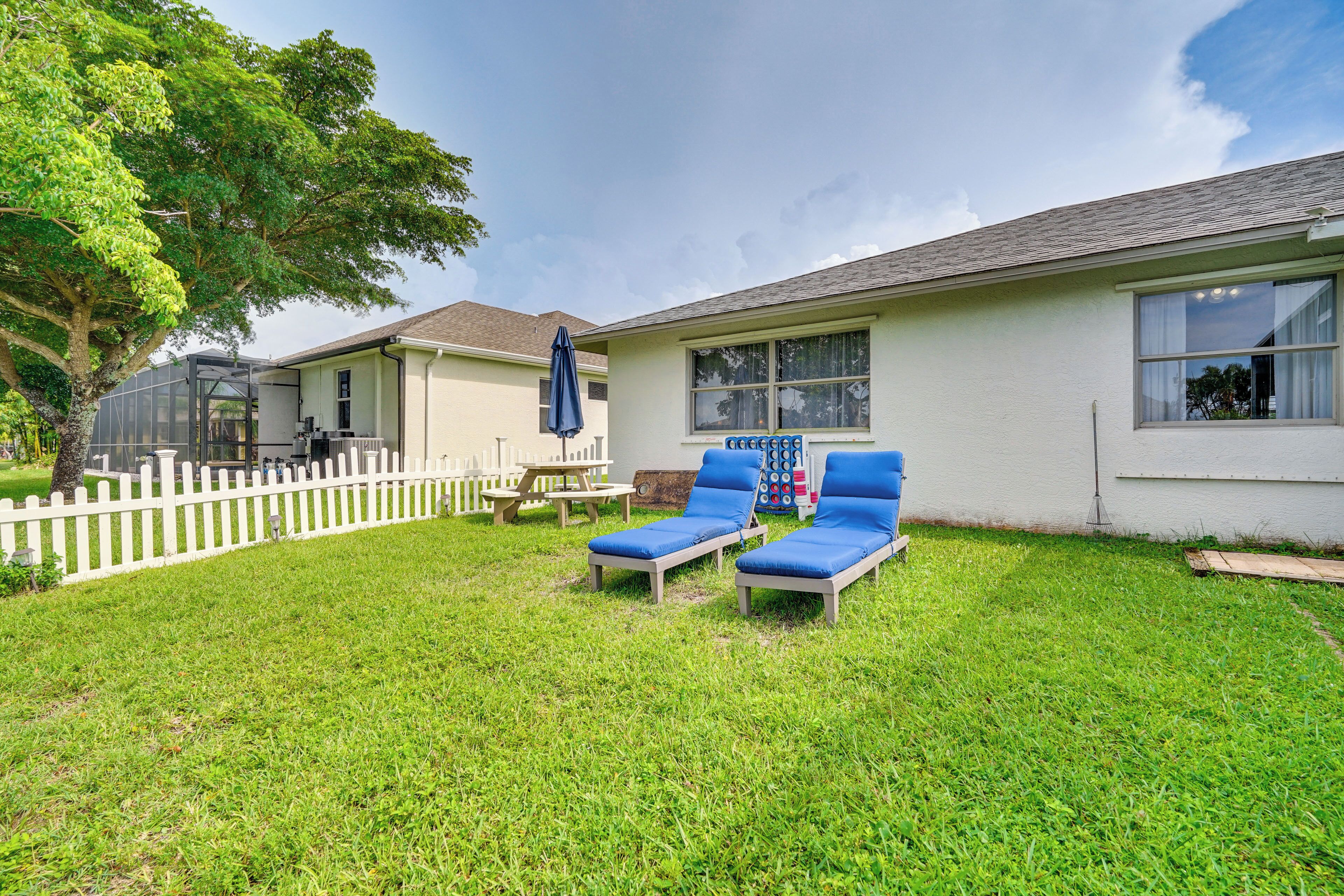 A house with a white exterior, a fenced backyard, two blue lounge chairs, a picnic table, and a large tree.