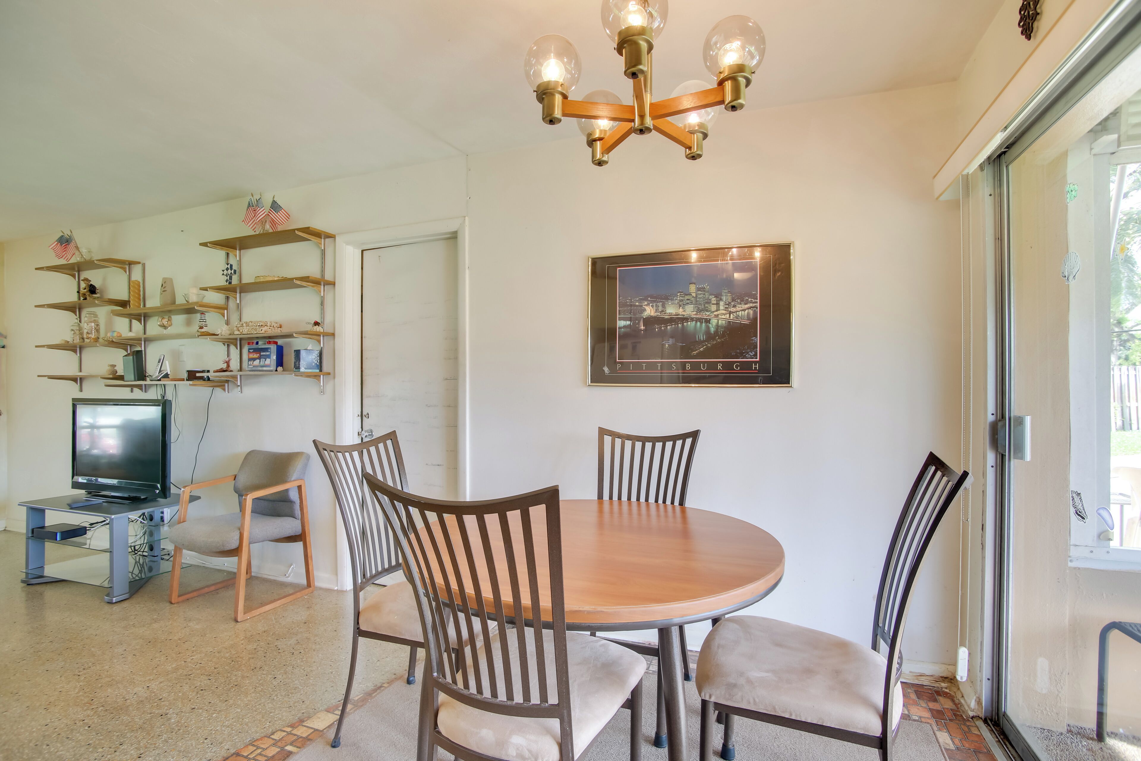 A dining area with a round wooden table, four chairs, a chandelier, a television on a stand, shelves with various items, and a framed picture of Pittsburgh on the wall.