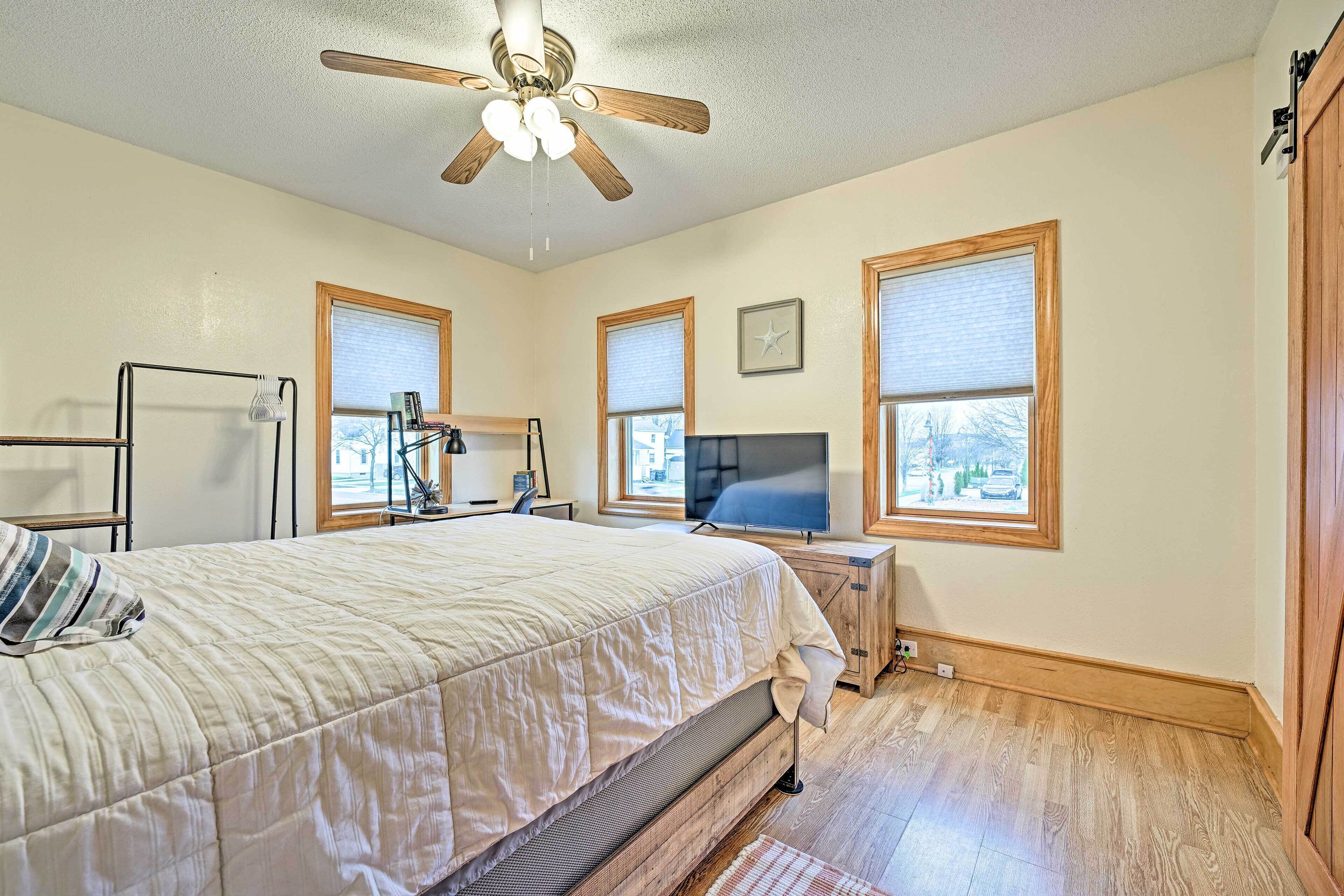A bedroom with a bed, a ceiling fan, a TV, and windows with blinds.