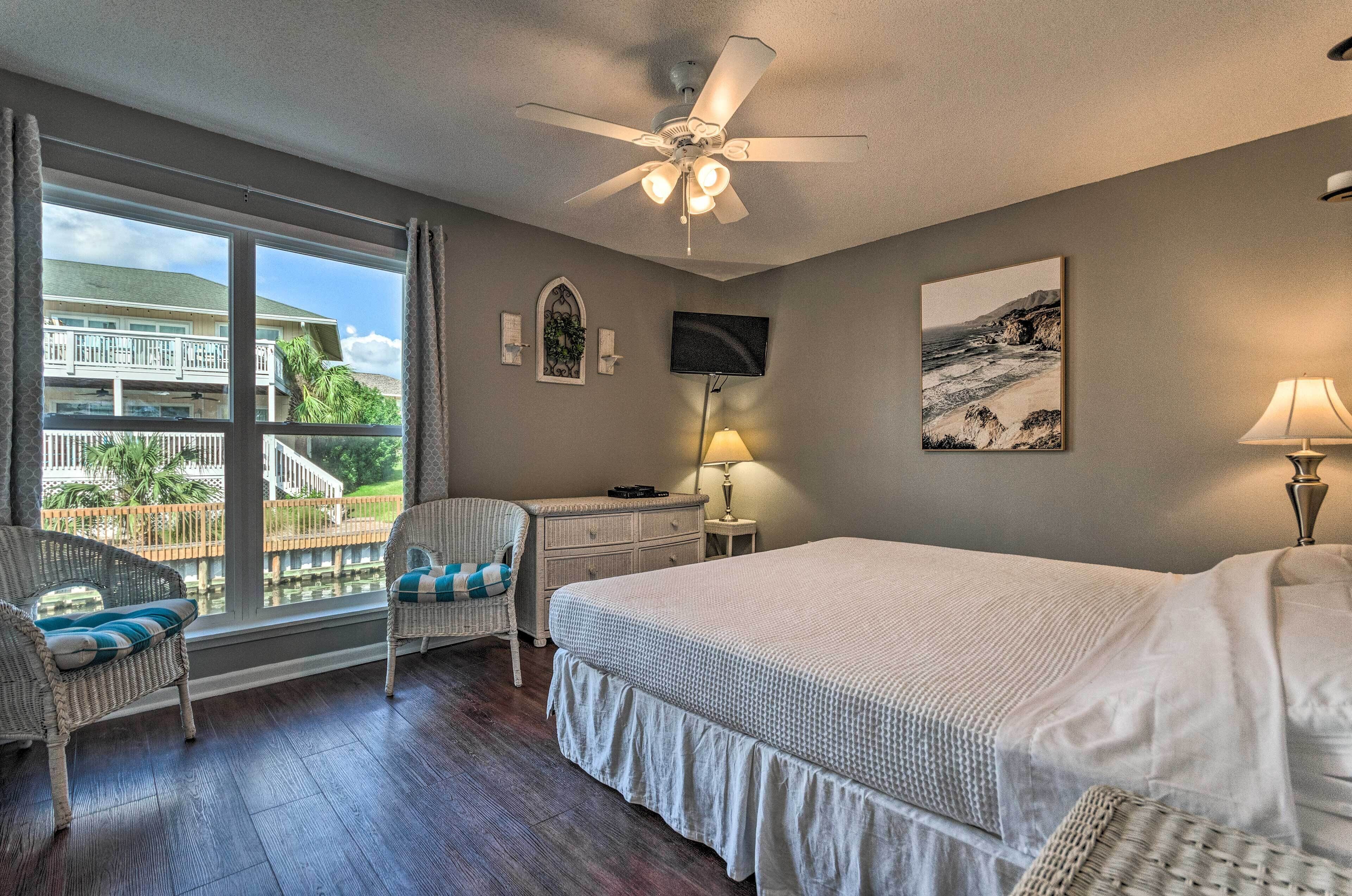 A bedroom with a large bed, a ceiling fan, a desk with a chair, a window with a view of a building and palm trees, and a lamp.