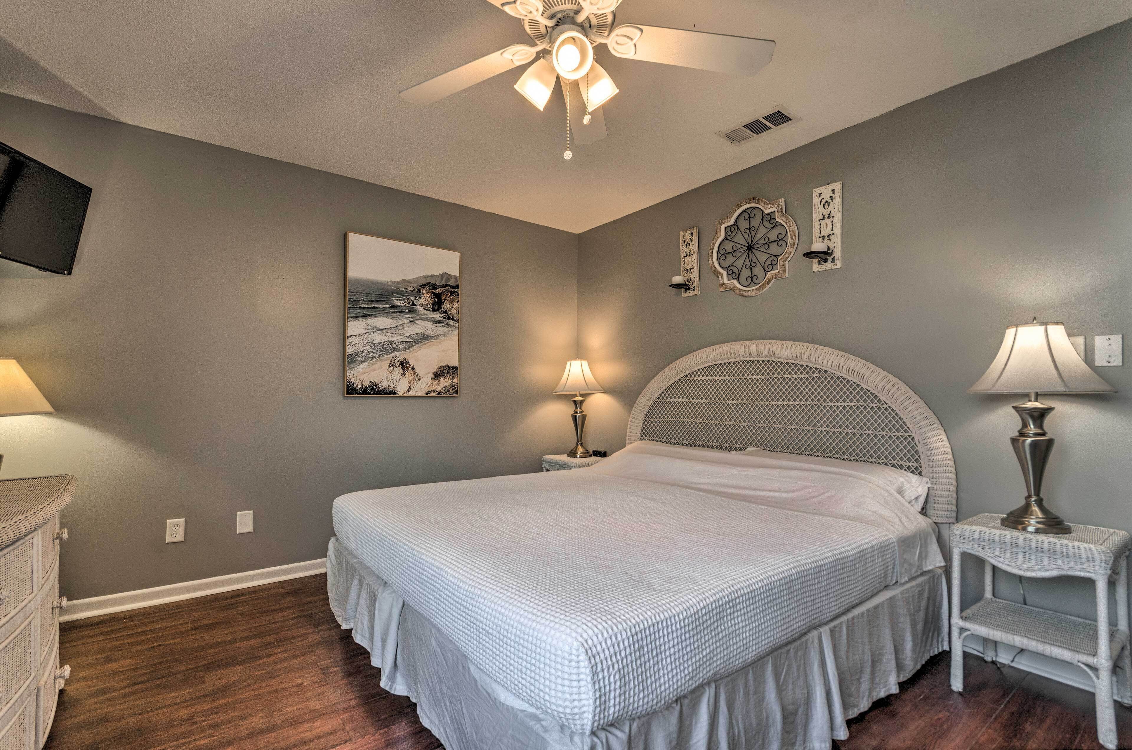 A bedroom with a large bed, two bedside tables with lamps, a ceiling fan, a wall-mounted TV, and a framed picture.