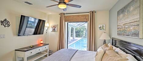 A bedroom with a bed, a desk, a television, a ceiling fan, and a view of a swimming pool.