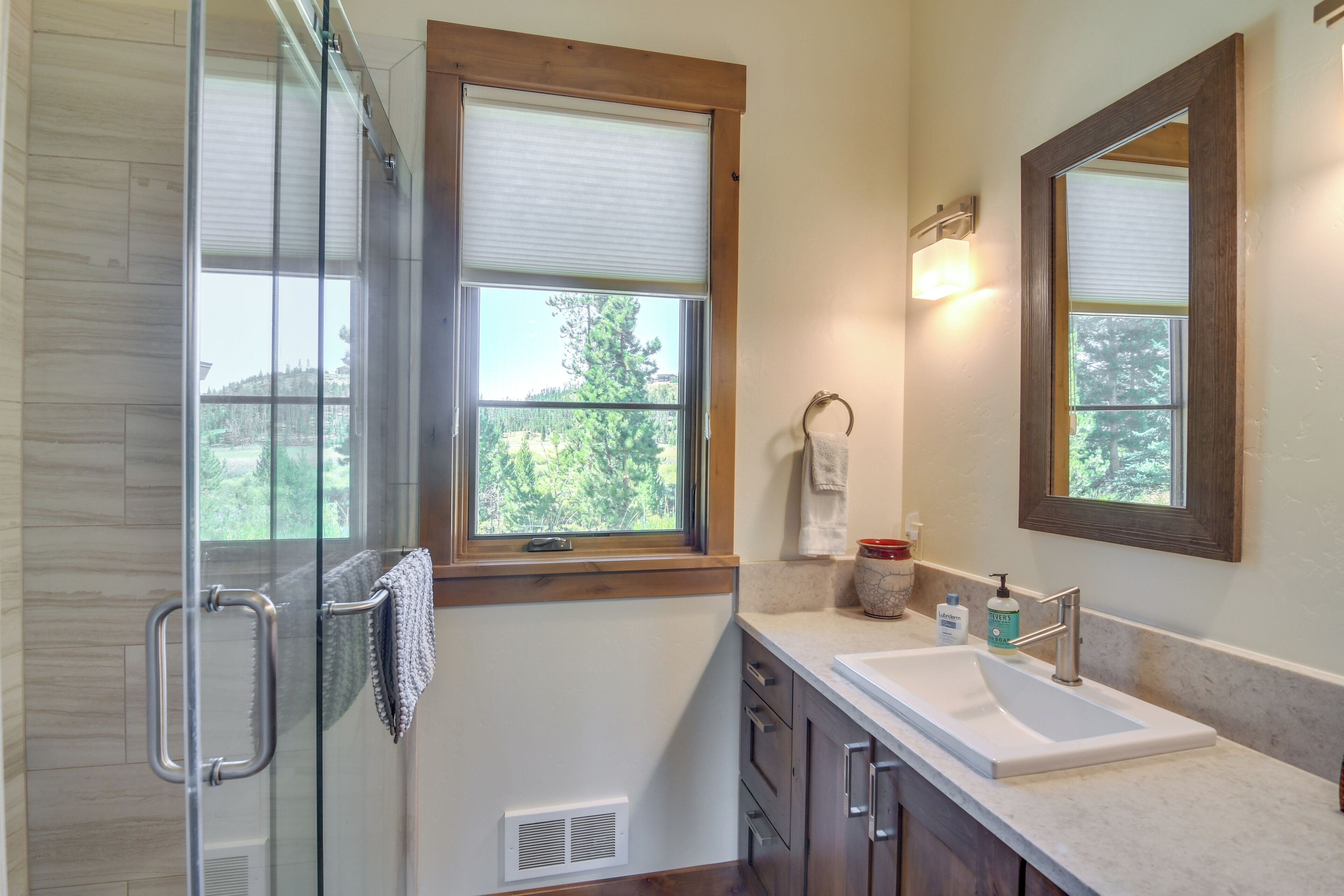 A modern bathroom with a glass shower enclosure, a large window with a view of greenery, a sink with a mirror, and a towel rack.