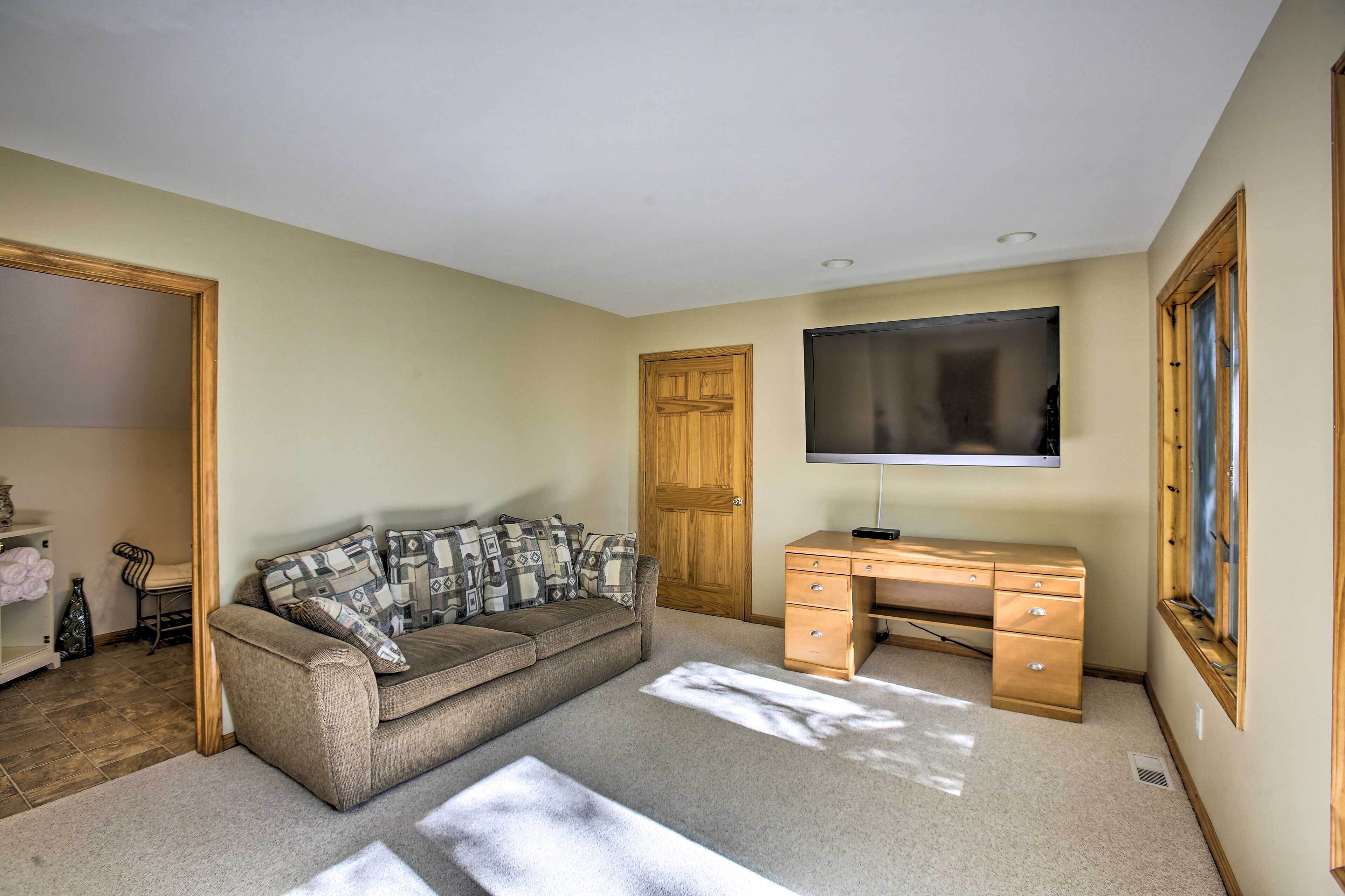 A room with a beige carpet, a brown sofa with patterned cushions, a wooden desk with drawers, a flat-screen TV mounted on the wall, and a wooden door.