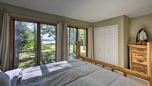 A bedroom with a large window, a bed, a wooden dresser, and a white door. - Interlochen Home on Green Lake w/ Dock & Beach! (Interlochen)