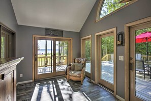 A room with a wicker chair, a wooden deck, and a view of a forest. - Interlochen Home on Green Lake w/ Dock & Beach! (Interlochen)