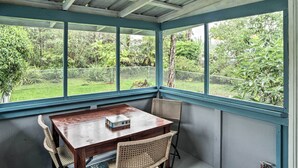 A covered patio with a wooden table and wicker chairs, large windows offering a view of a lush garden. - P'hoa Home 4 Mi to Kilauea Caverns of Fire! (Pahoa)