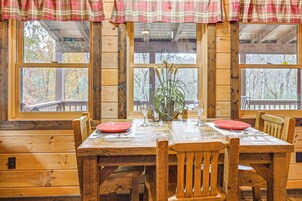 Una mesa de comedor de madera con dos platos rojos, rodeada de sillas de madera, frente a paredes de madera y ventanas grandes con vistas a árboles.