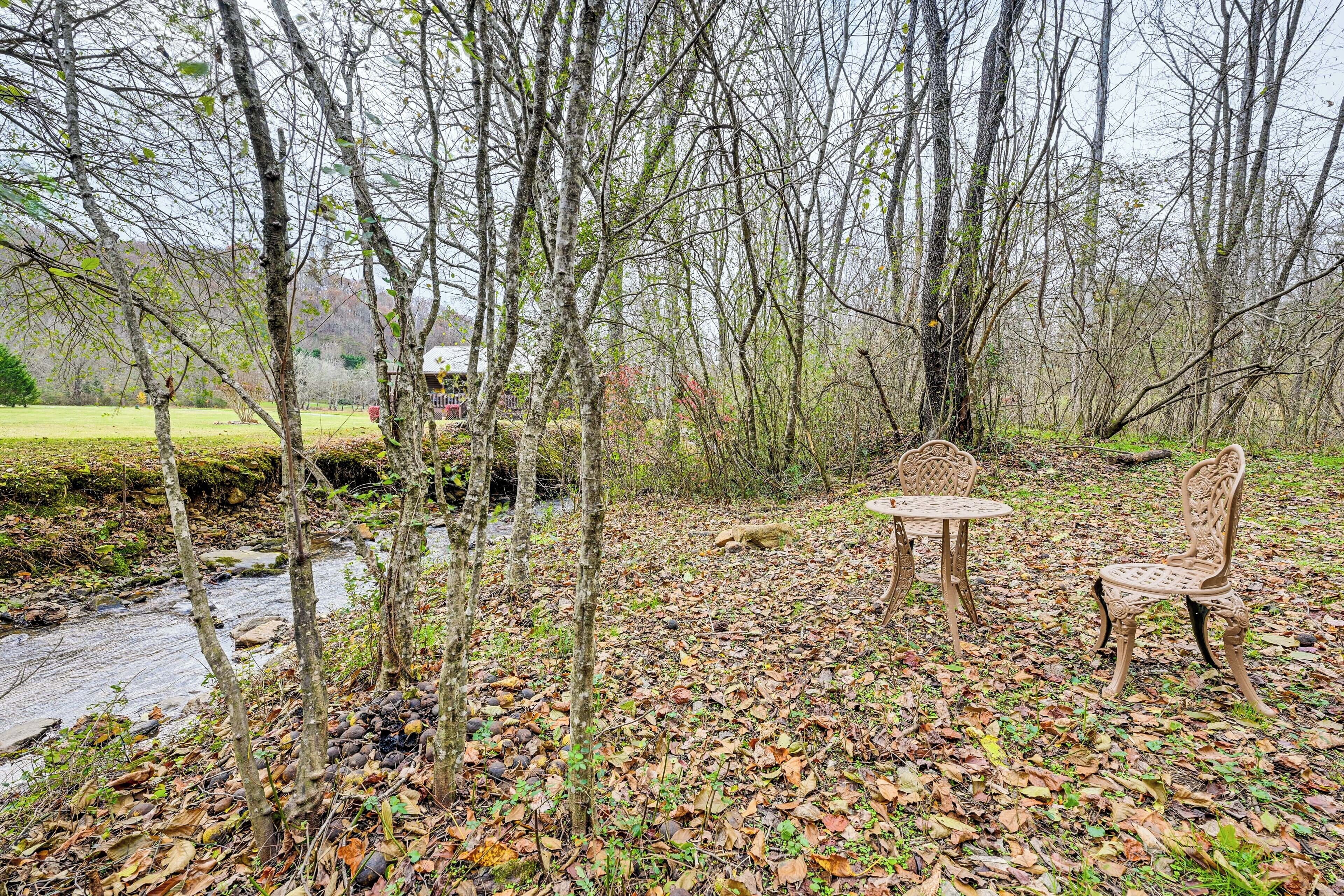 A rustic outdoor seating area with a table and two chairs, surrounded by trees and a stream.