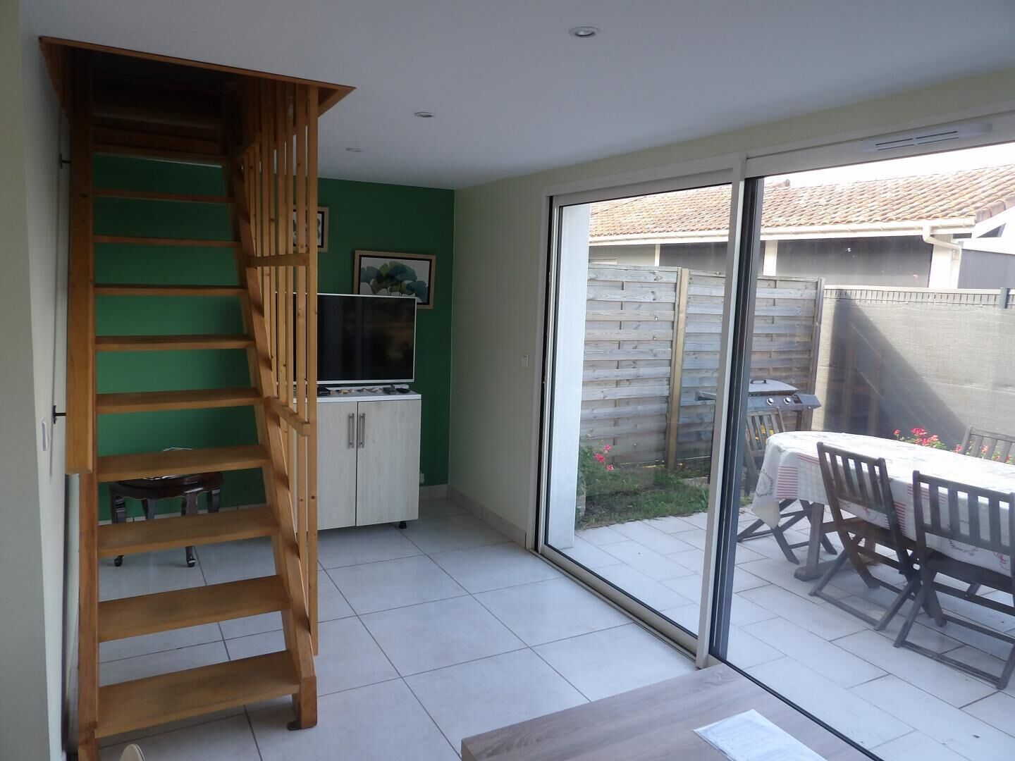 Una habitación con escalera de madera, un televisor sobre un mueble, una mesa de comedor con sillas y un ventanal corredero que da a un espacio exterior.