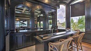 A modern kitchen with dark cabinetry, a central island with bar stools, and large windows offering a view of palm trees. - Luxe Cape Coral Escape w/ Boat Dock + Cabana! (Cape Coral)