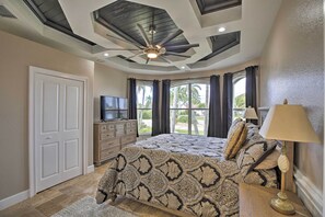 Ein Schlafzimmer mit Deckenventilator, einem Bett mit gemusterten Betttextilien, einem Fernseher und Blick auf Palmen durch große Fenster.