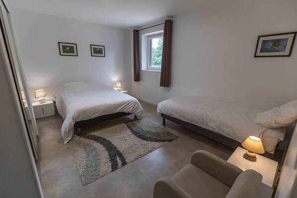 Un dormitorio con una cama doble, una cama individual, una silla, una mesita de noche, una lámpara y dos cuadros en la pared.
