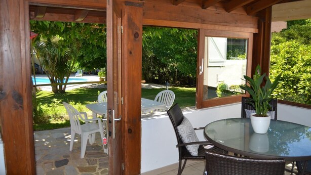 Un patio al aire libre con una mesa de vidrio y sillas de mimbre, rodeado de vigas de madera y vistas a una piscina y follaje.
