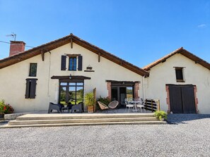 Edificio de dos pisos con camino de grava, puerta principal y zona de patio con mobiliario exterior.