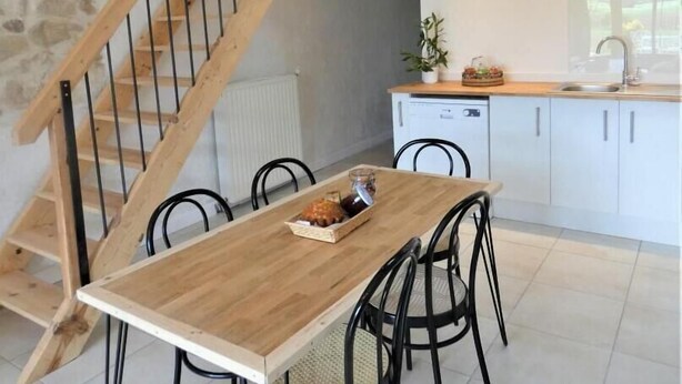 A dining area with a wooden table and chairs, a kitchen with white cabinets, and a wooden staircase. - Le Petit Saou du Becq (SAINT LON LES MINES)