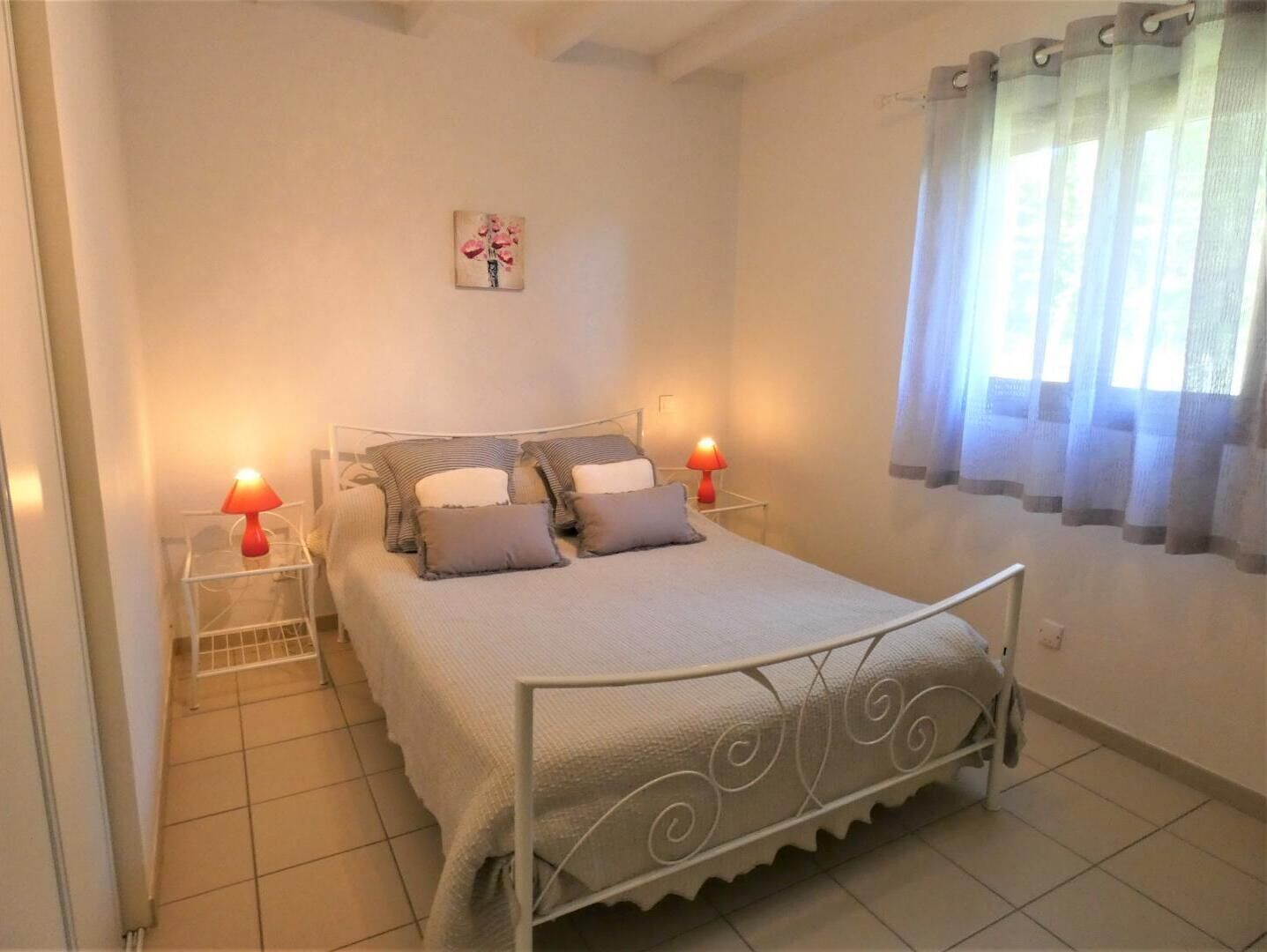 A neatly arranged bedroom with a bed, two bedside lamps, a wall-mounted artwork, and a window with sheer curtains.