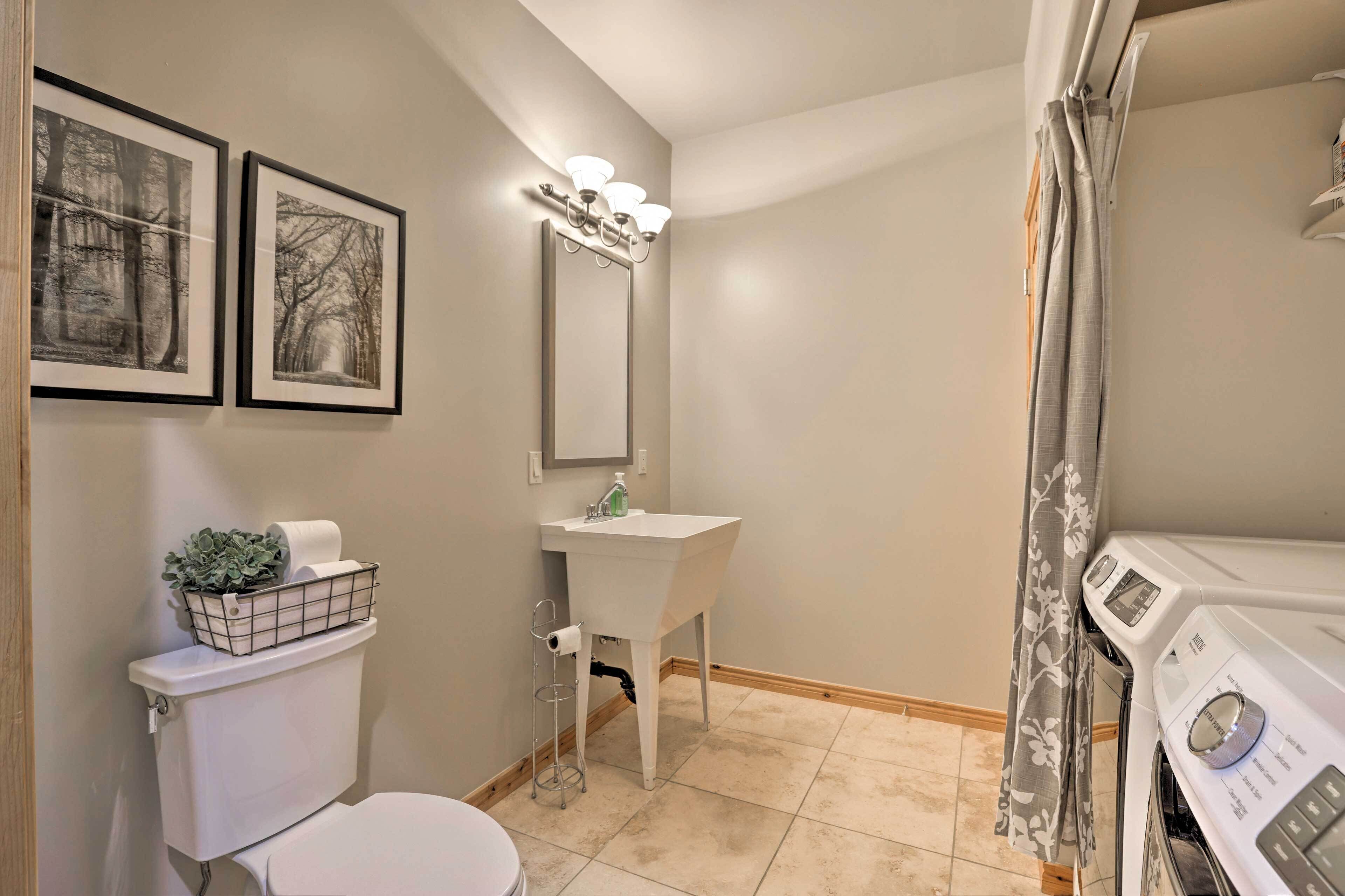 A bathroom with a toilet, sink, and washing machine. There are two framed pictures on the wall, a light fixture above the sink, and a curtain on the window.