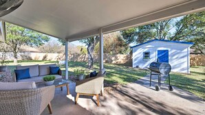 Une terrasse couverte, meublée de mobilier en osier, avec un barbecue et une petite maison en arrière-plan.