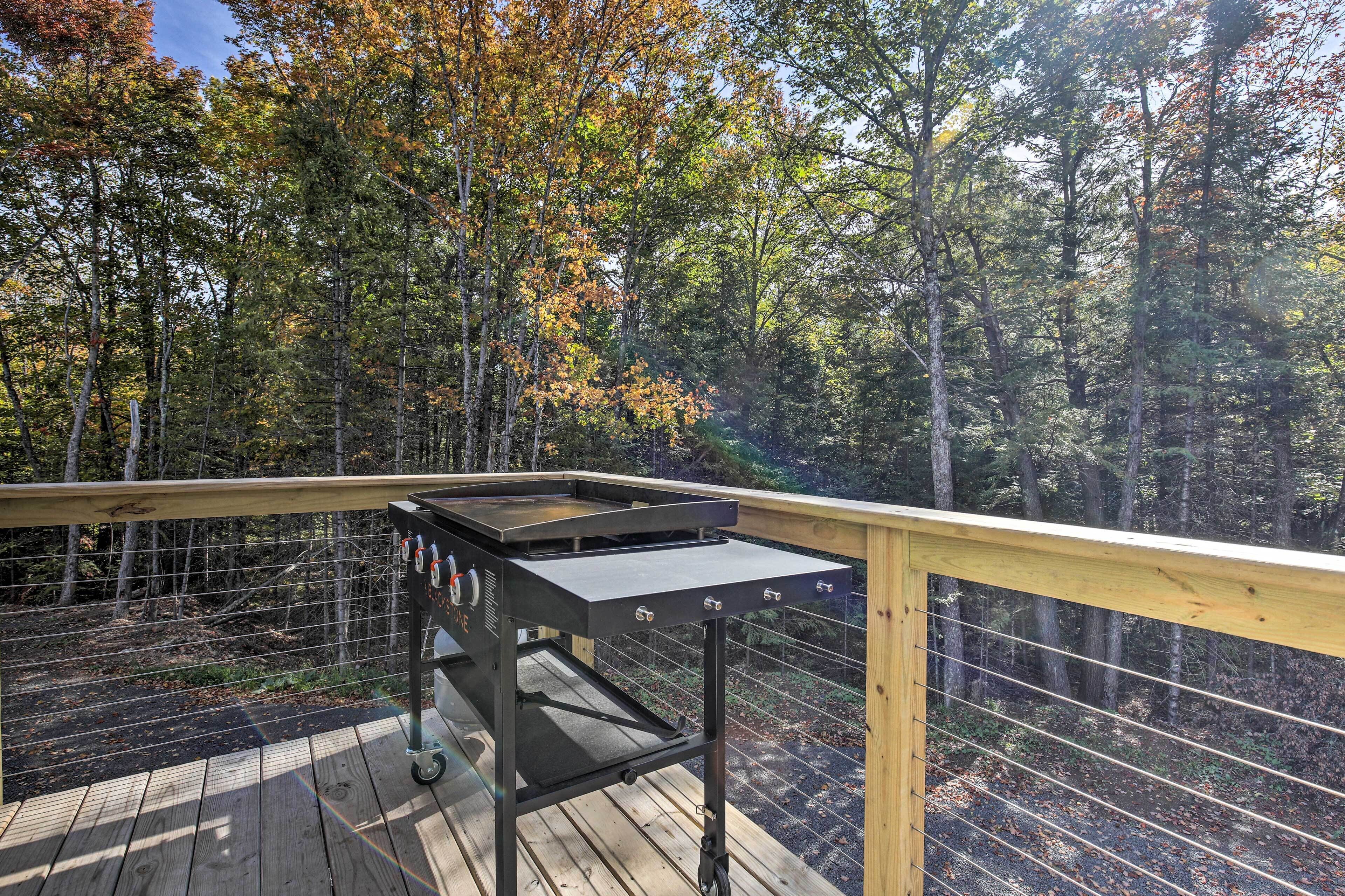 A deck with a barbecue grill, surrounded by a wooden railing and overlooking a forested area.