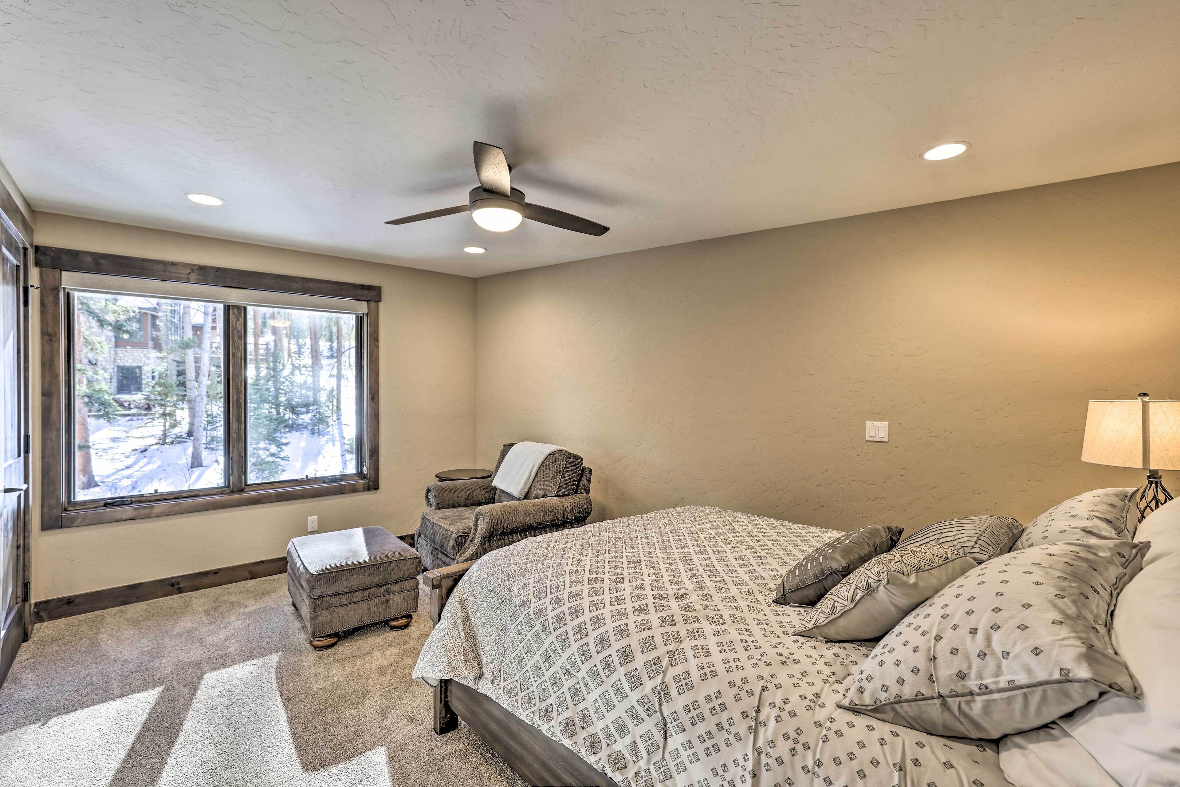 A bedroom with a large bed, a chair, a small ottoman, a ceiling fan, and a window with a view of trees.