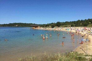 A beach with clear water, people swimming and sunbathing, and a small building in the distance. - Arbousiers - Morcenx (MORCENX)