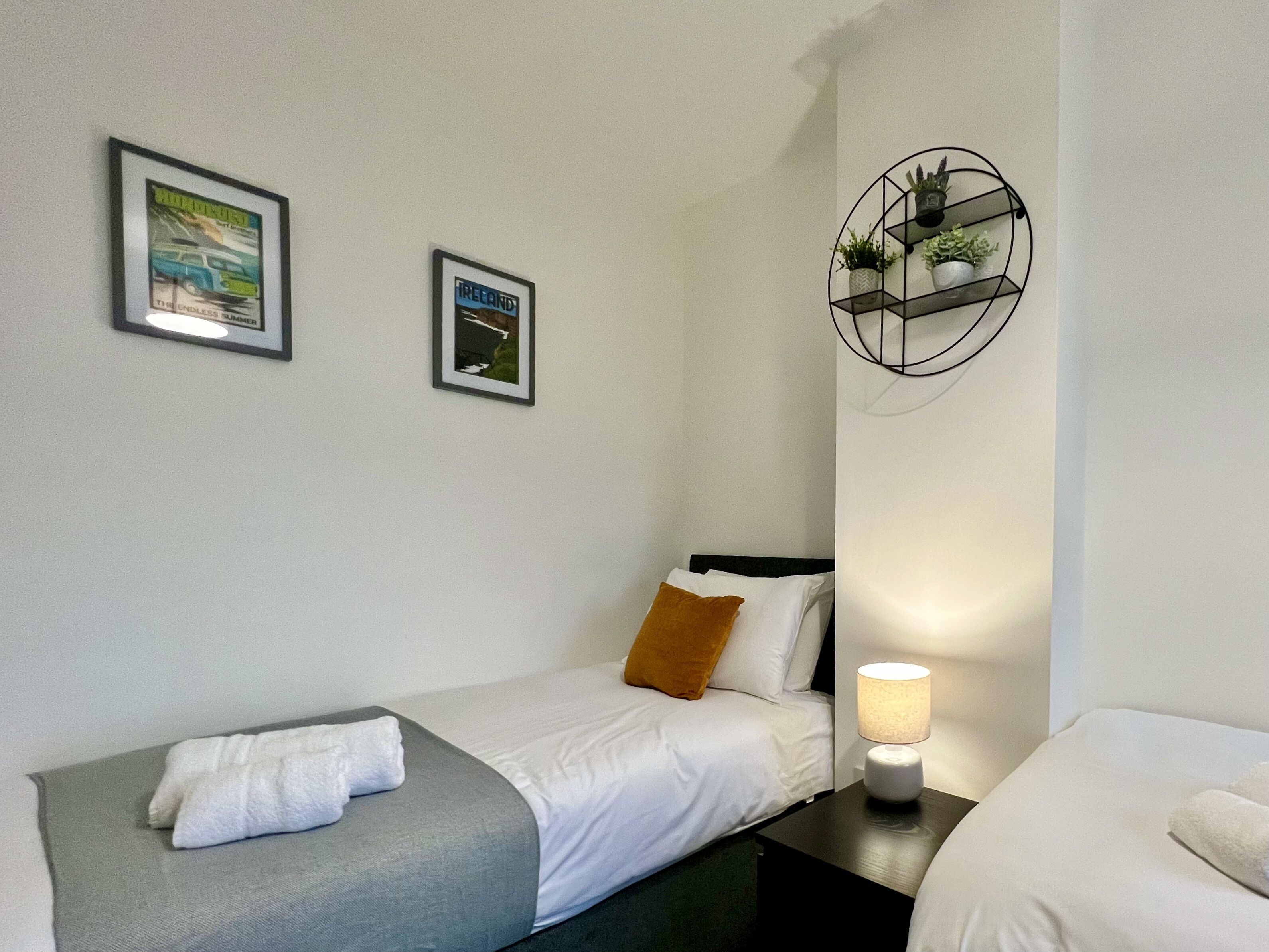 A hotel room with two single beds, a nightstand with a lamp, a wall-mounted shelf with plants, and two framed pictures.