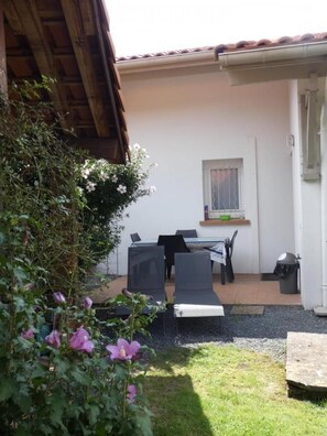 Un patio con una mesa y sillas, una ventana con cortina y un pequeño jardín lleno de flores.