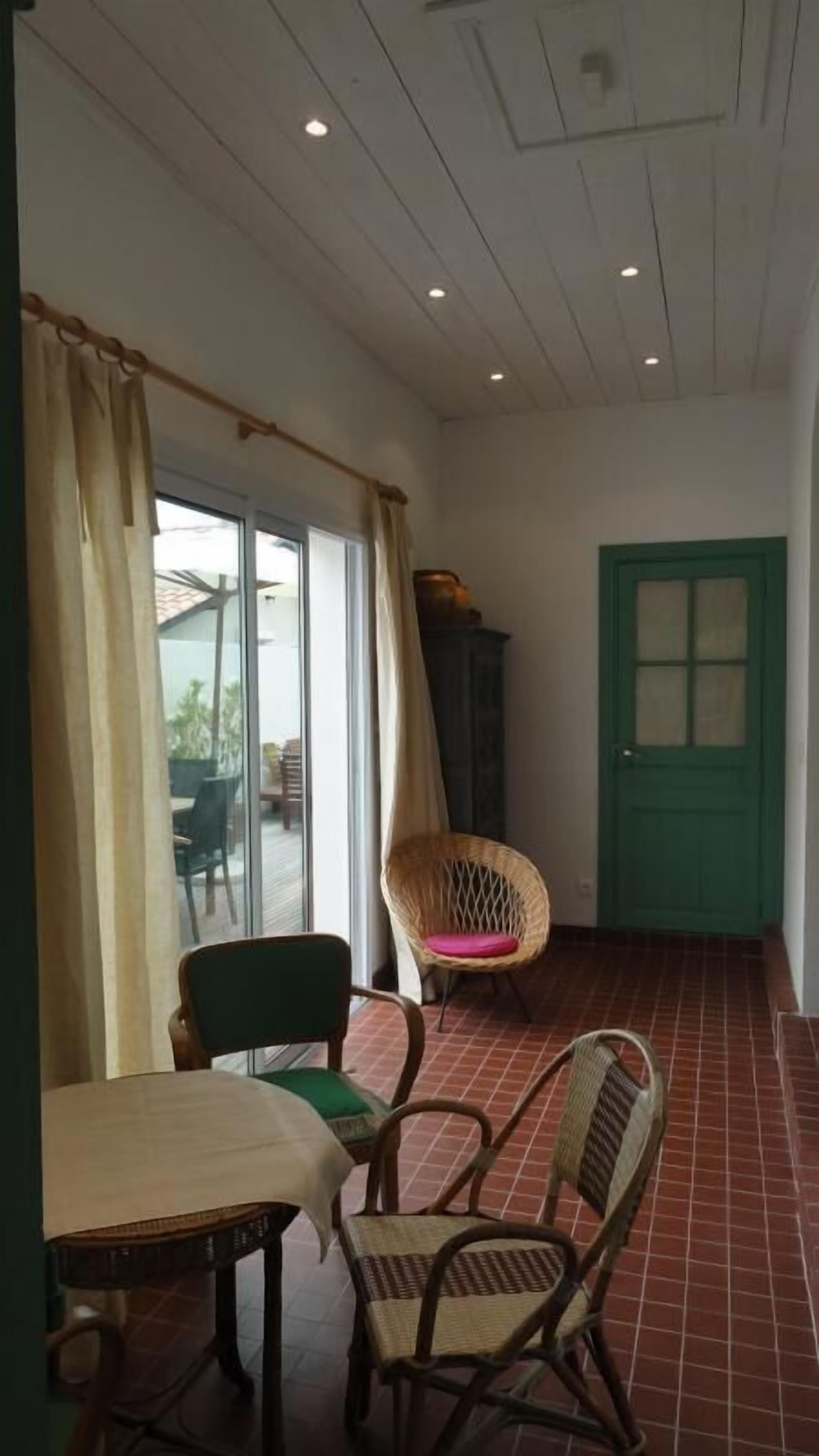 A room with a table and chairs, a wicker chair with a cushion, a green door, and a window with curtains.