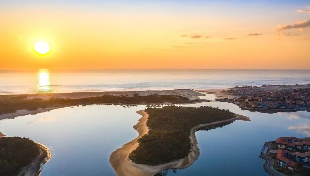 An aerial view of a coastal area with a sunset, calm waters, and a small island with buildings.
