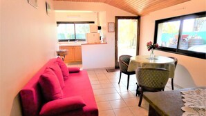 A living room with a red sofa, a dining table with chairs, a kitchen in the background, and a window with a view of the outside. - L'ecureuil - Soustons (SOUSTONS)