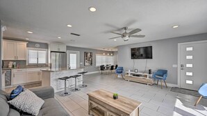 A modern living room with a grey sofa, a coffee table, and a TV. There is a kitchen area with white cabinets and a refrigerator. - Remodeled Naples Getaway w/ Pool ~ 1 Mi to Beach! (Naples)